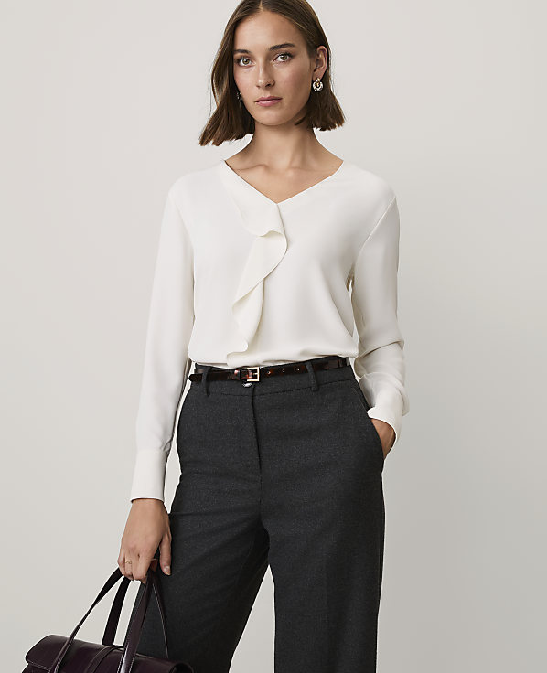 A woman wearing a white ruffled blouse and dark grey trousers stands against a plain light grey background.