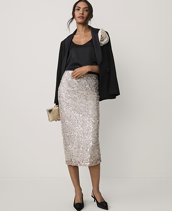 A woman wearing a black blazer and a shimmering silver sequined skirt stands against a plain, light-colored background.