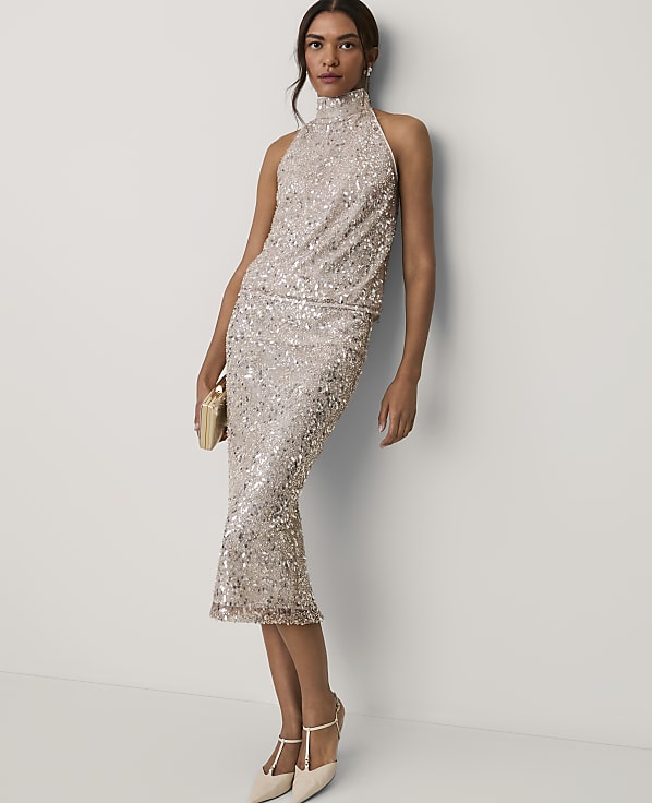 A woman wearing a shimmering champagne-colored sequin midi dress and heels stands against a plain white wall.