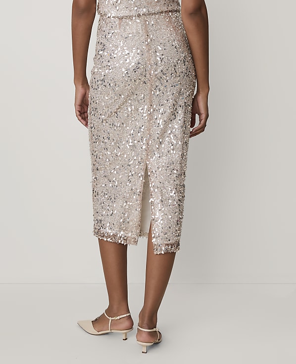A person wears a shimmering gold sequined midi skirt and beige heels against a plain, light-colored background.