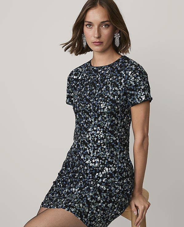 A woman wearing a sparkling blue and black sequined dress sits on a wooden chair against a plain light gray background.