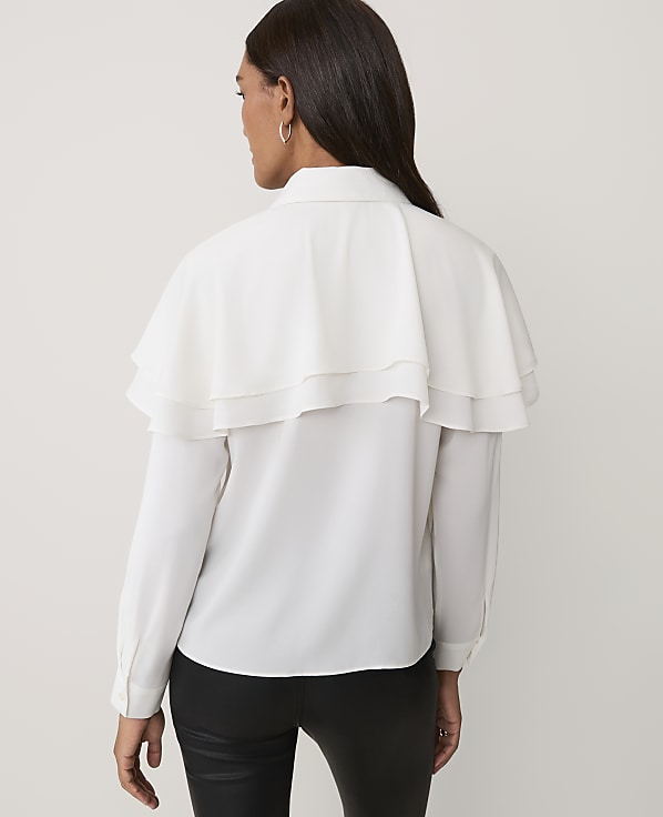 A woman wearing a white ruffled blouse and black pants stands against a plain light-colored background.