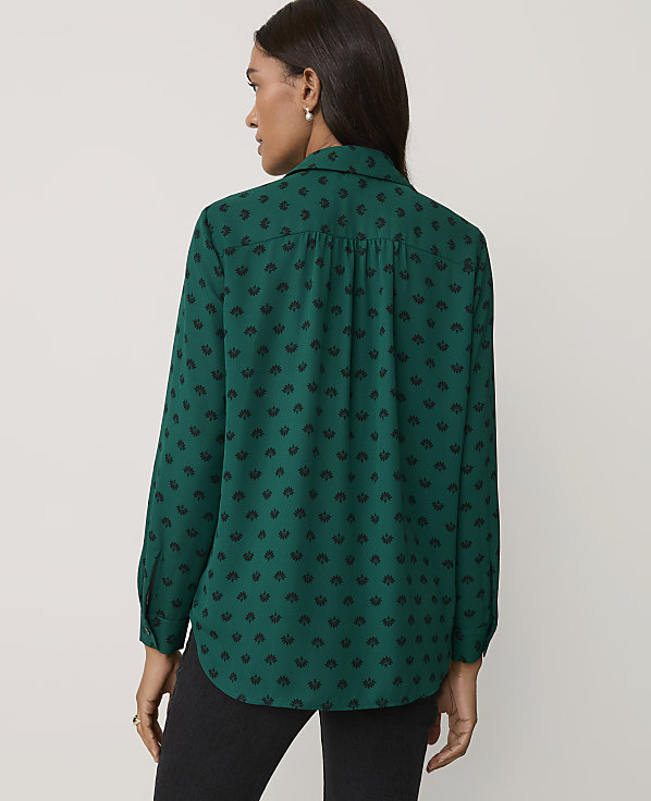 A woman wearing a green patterned shirt and black trousers stands against a plain, light-colored background.