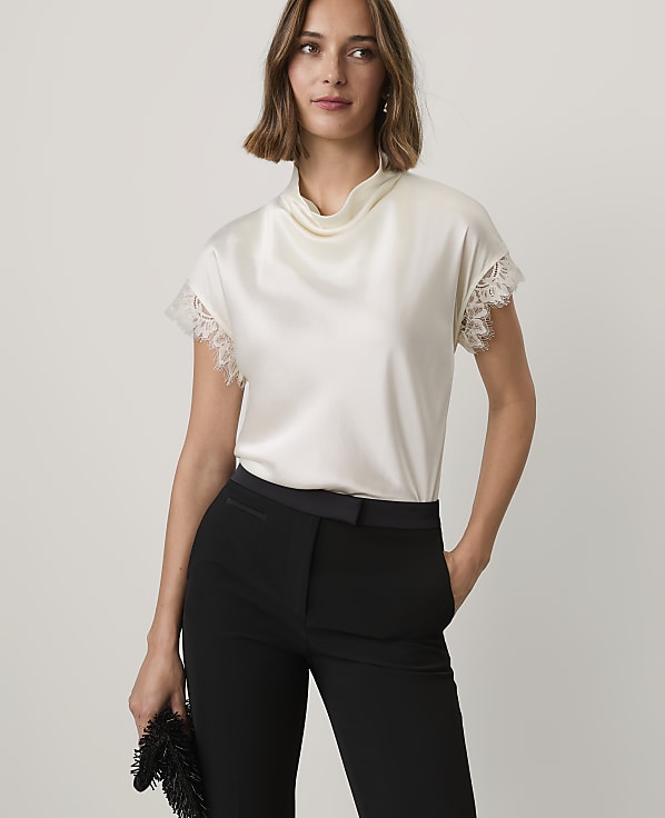 A woman wearing a white satin top and black trousers stands against a plain, light-colored background.