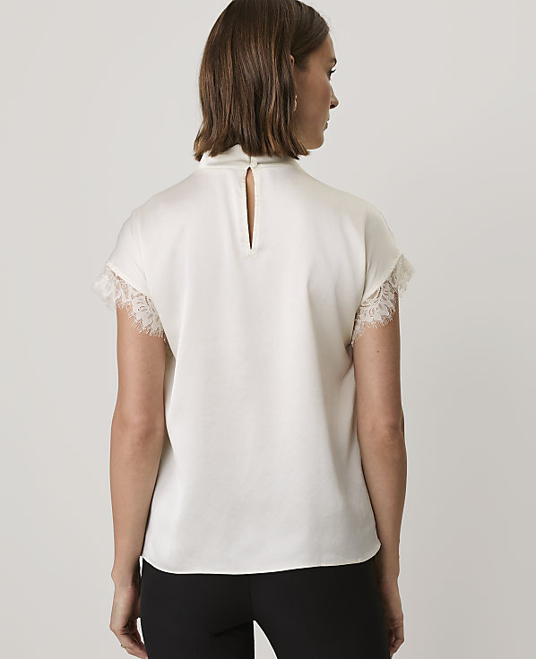 A woman wearing a white lace-trimmed top and black trousers stands with her back to the camera against a plain white background.