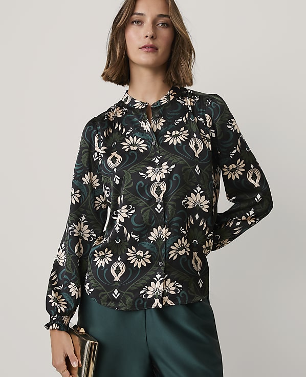 A woman wearing a floral print blouse and dark green trousers stands against a plain, light-colored background.