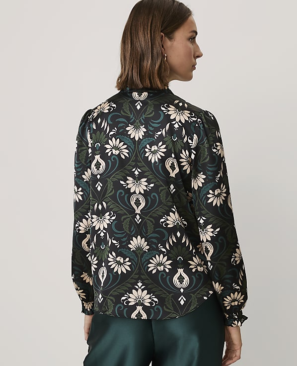 A woman wearing a floral print blouse and dark green satin trousers stands against a plain light gray background.