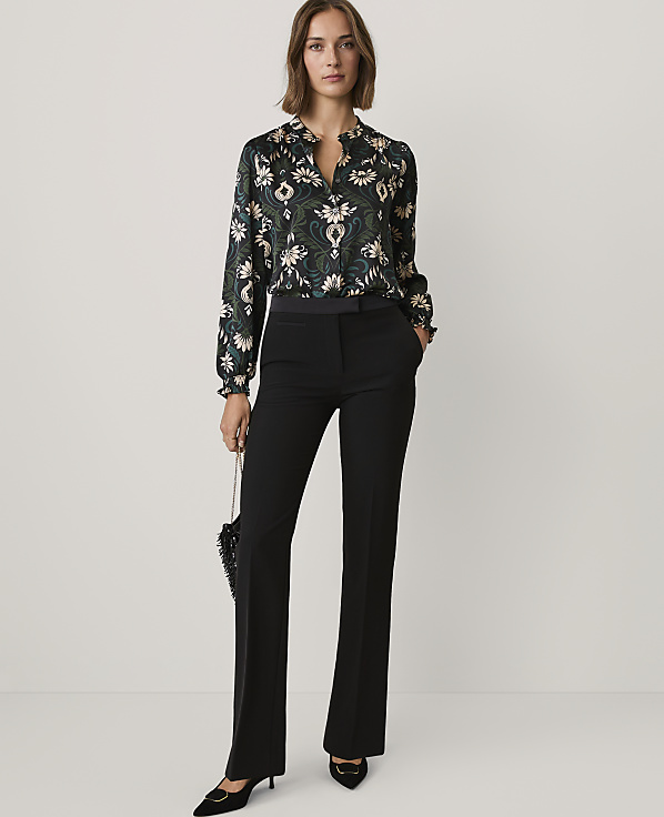 A woman wearing a patterned blouse and black trousers stands against a plain light grey background.