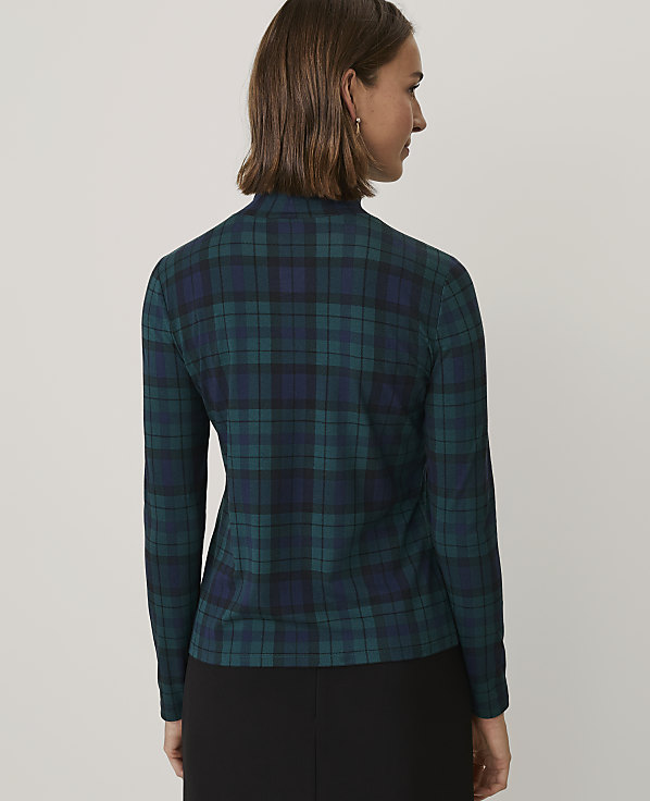 A woman wearing a green and navy blue plaid top and black pants stands against a plain light gray background.