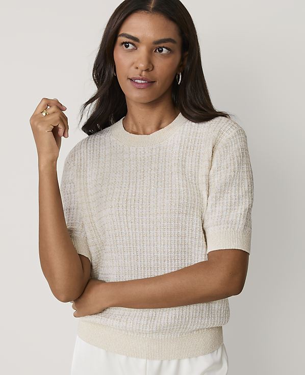 A woman wearing a cream-colored textured short-sleeve sweater and white pants stands against a plain, light-colored background.