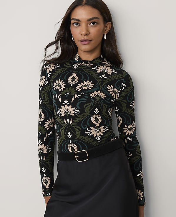 A woman wearing a floral print long-sleeved top and a black skirt stands against a plain white background.