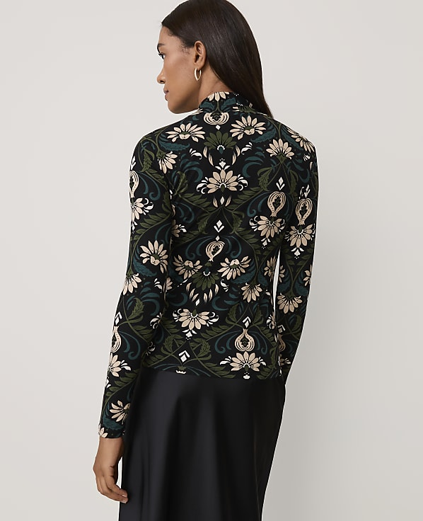 A woman wearing a floral patterned top and a black skirt stands against a plain light gray background.