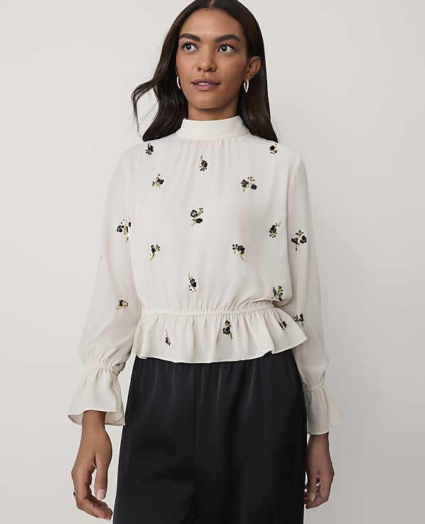 A woman wearing a white floral-embroidered blouse and black trousers stands against a plain light-colored background.