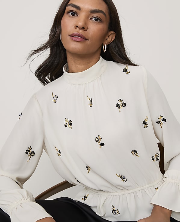 A woman wearing a white floral-embroidered blouse and black pants sits against a plain, light-colored background.
