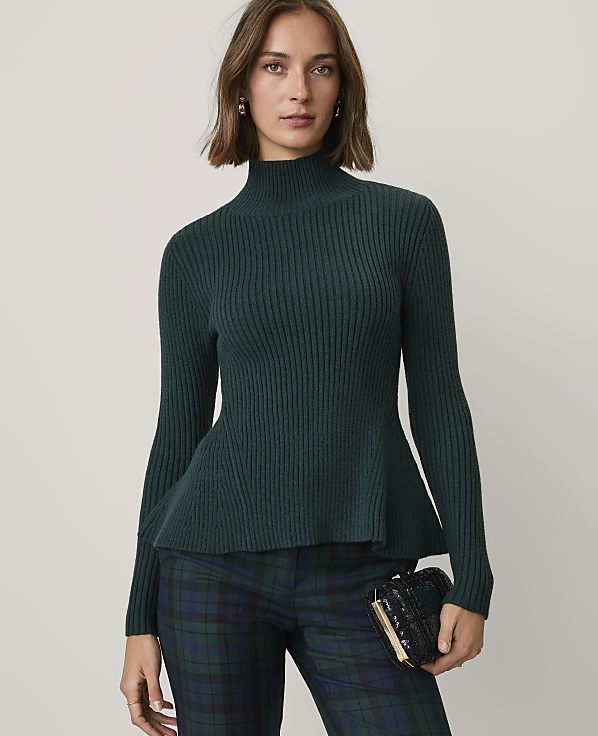 A woman wearing a dark green ribbed peplum sweater and plaid trousers stands against a plain light gray background.