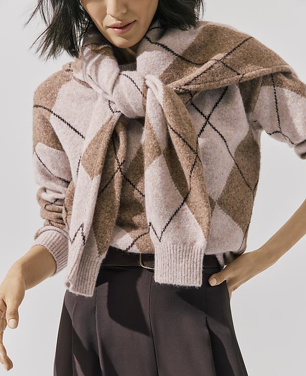 A person wears a pink and brown argyle sweater with a matching cardigan tied around their shoulders against a plain white background.