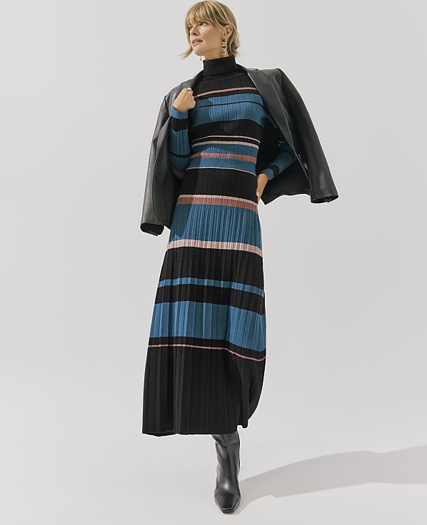 A woman wearing a striped blue and black pleated dress with a leather jacket draped over her shoulders stands against a plain white background.