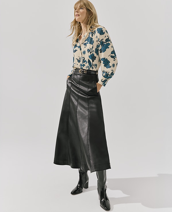 A blonde woman wearing a floral blouse and a long black leather skirt stands against a plain white background.