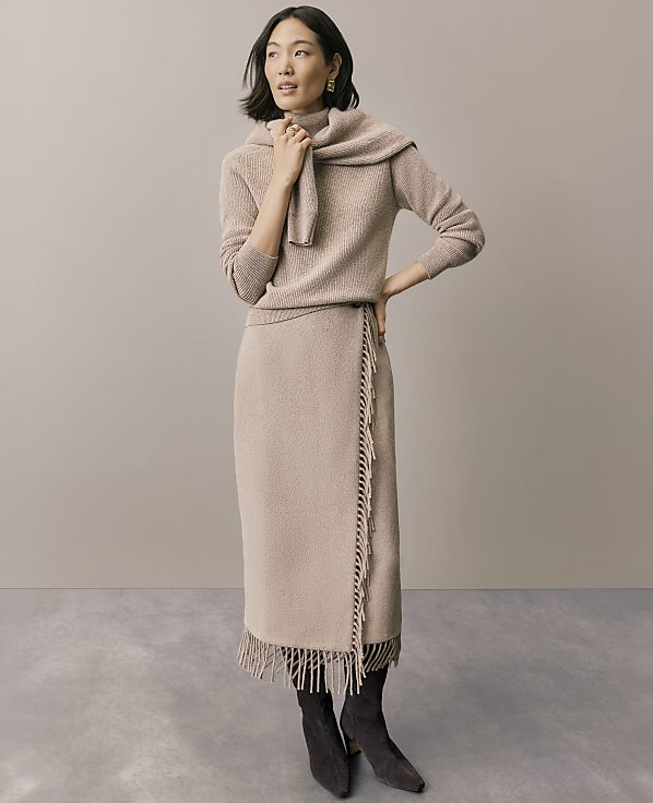 A woman wearing a beige knit sweater and matching fringed skirt stands against a plain, neutral-toned wall and floor.