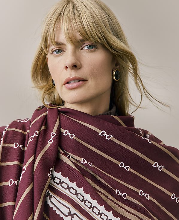 A blonde woman wearing a patterned burgundy and beige scarf and gold earrings poses against a plain, neutral background.