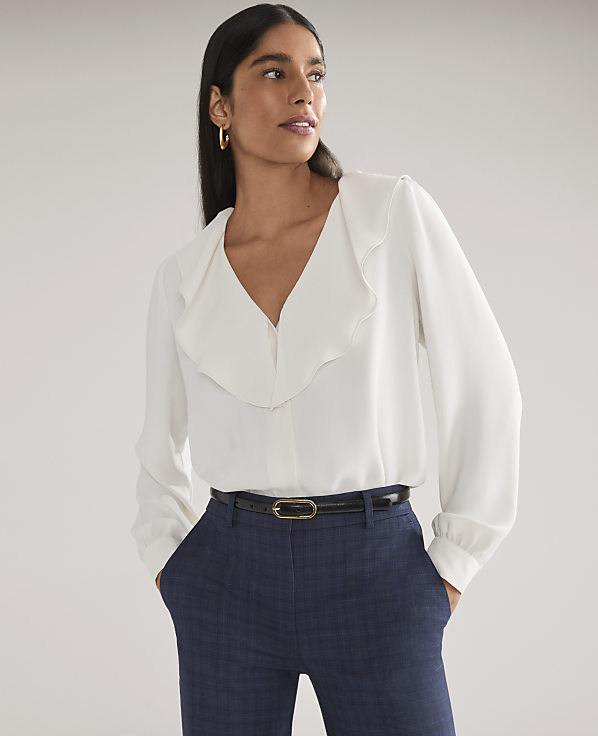 A woman wearing a white ruffled blouse and navy blue trousers stands against a plain light gray background.