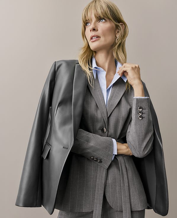A blonde woman wearing a pinstriped grey suit and light blue shirt with a dark grey leather jacket draped over her shoulders stands against a plain beige background.