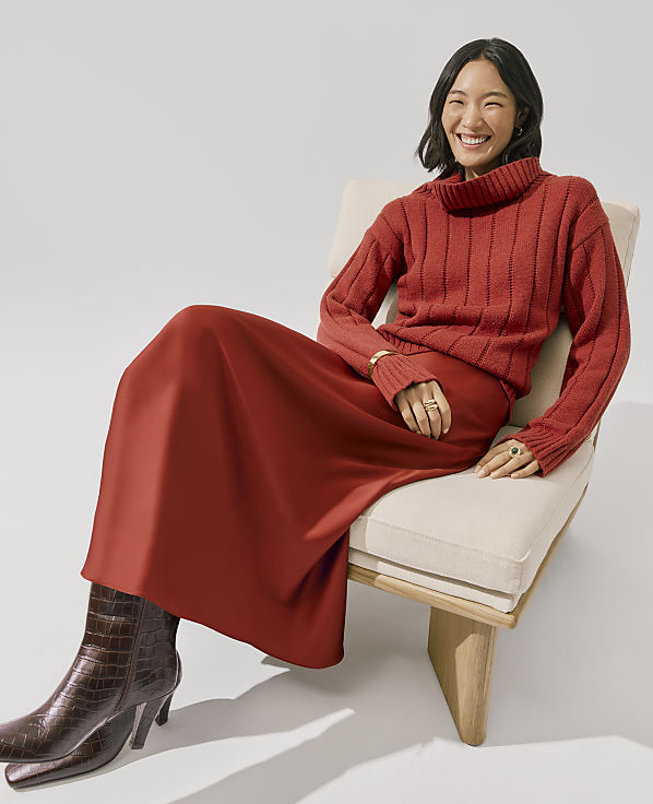 A smiling woman wearing a red ribbed sweater and a red satin skirt sits in a light-colored chair against a plain white background.
