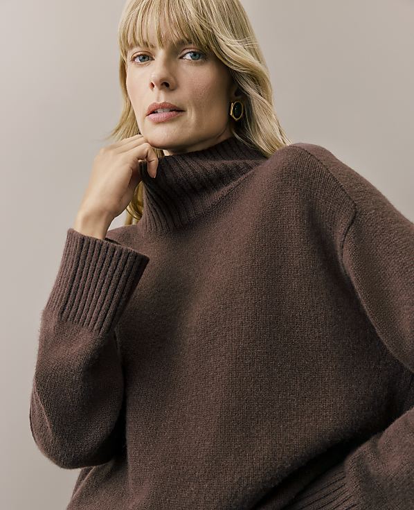A blonde woman wearing a brown turtleneck sweater poses against a plain, neutral-colored background.