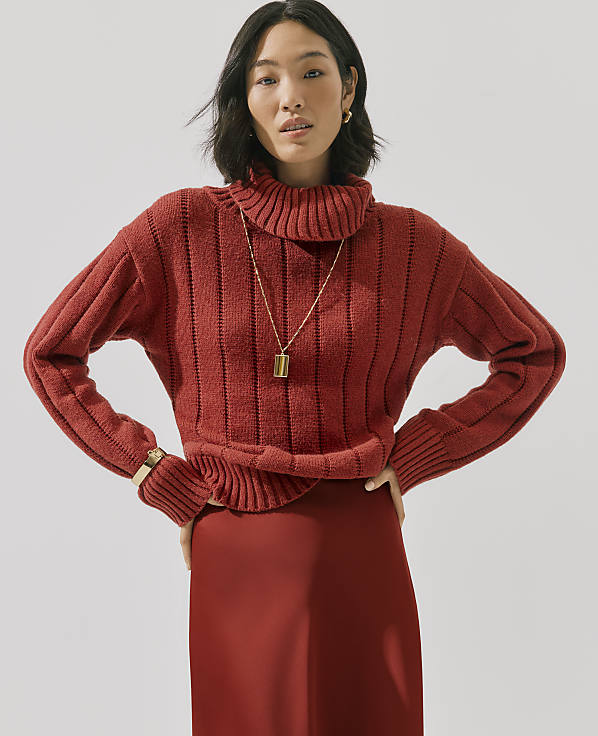 A woman wearing a red ribbed turtleneck sweater and matching skirt stands against a plain light gray background.