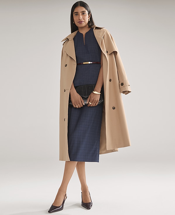 A woman wearing a navy blue dress and a tan trench coat stands against a plain light gray background.