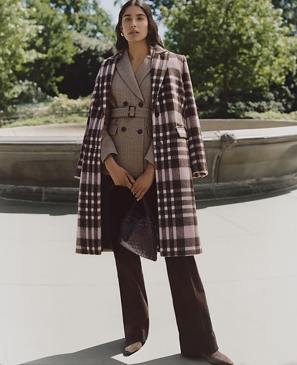 A woman wearing a plaid coat and brown trousers stands in the foreground before a stone fountain and lush green trees in the background.