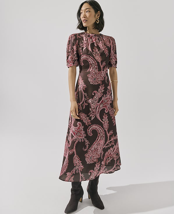 A woman wearing a brown and pink paisley print dress and dark boots stands against a plain white background.