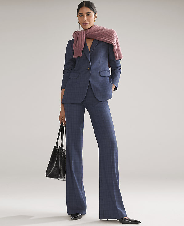 A woman wearing a blue plaid suit with a pink sweater draped over her shoulders and holding a black handbag stands against a plain light gray background.