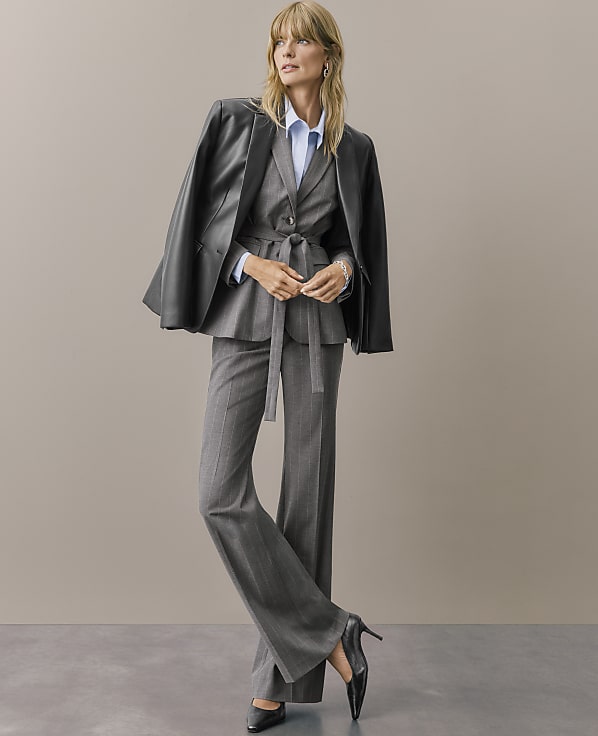 A woman wearing a grey pinstripe suit and a black leather jacket draped over her shoulders stands against a plain, neutral-toned background.