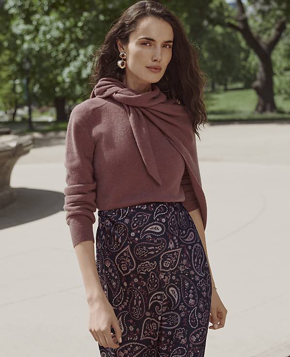 A woman wearing a mauve sweater and paisley trousers stands in the foreground against a blurred background of green trees and a paved walkway.