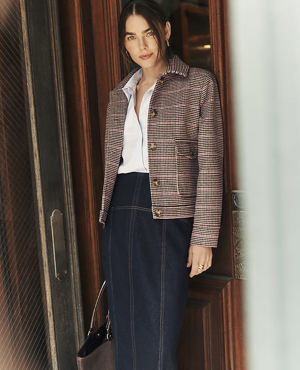 A woman wearing a patterned jacket, white shirt, and dark denim skirt stands in a wooden doorway.