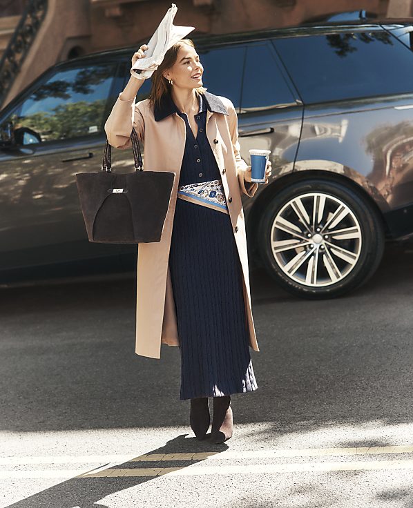 A woman in a tan coat and navy dress holds a coffee cup and a white hat, standing in front of a dark grey SUV.