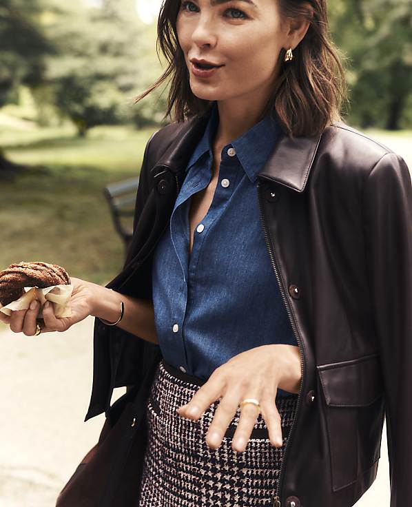 A woman wearing a leather jacket and denim shirt holds a cookie in the foreground against a blurred park background.