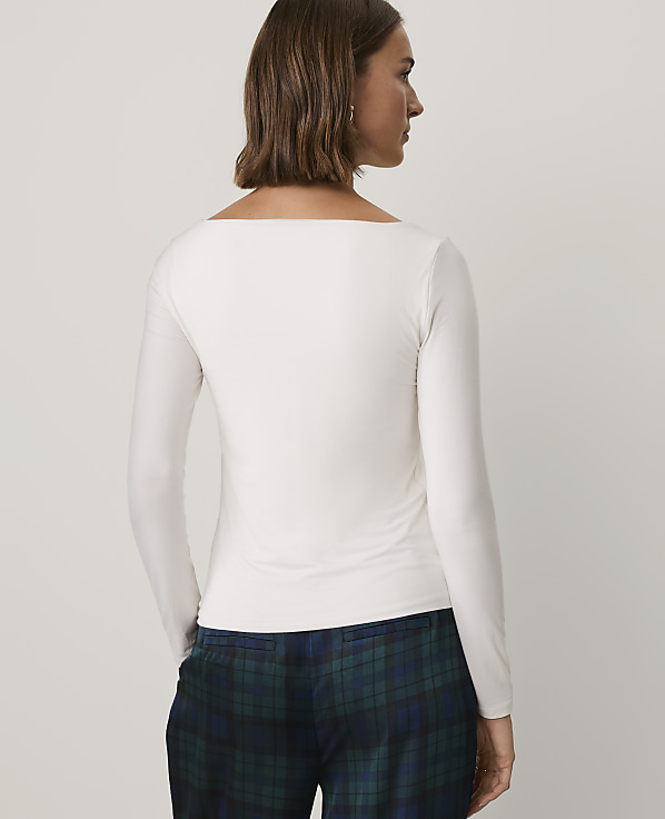 A woman wearing a white long-sleeved top and blue plaid trousers stands against a plain light gray background.