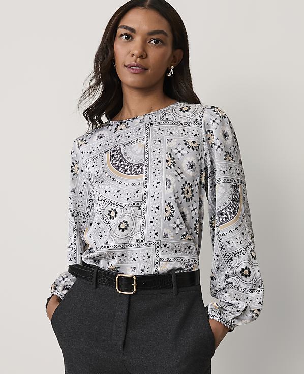 A woman wearing a patterned grey blouse and dark trousers stands against a plain white background.