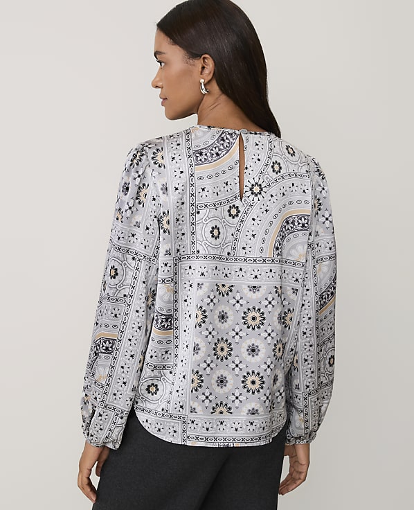 A woman wearing a patterned grey and white blouse and dark trousers stands against a plain light-colored background.