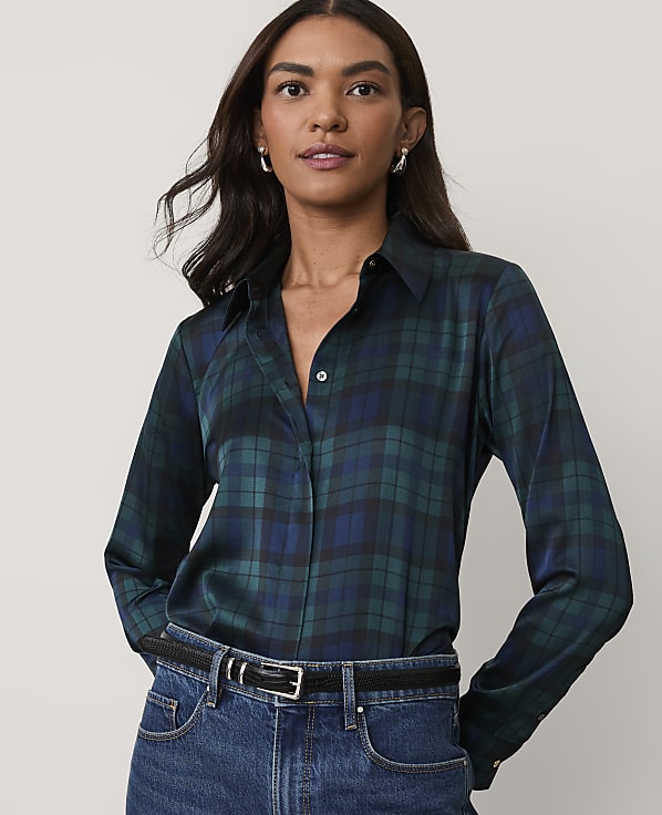 A woman wearing a dark green plaid button-down shirt and blue jeans stands against a plain, light-colored background.