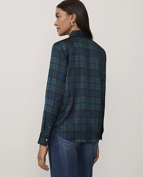 A woman wearing a green and blue plaid shirt and blue jeans stands against a plain light gray background.