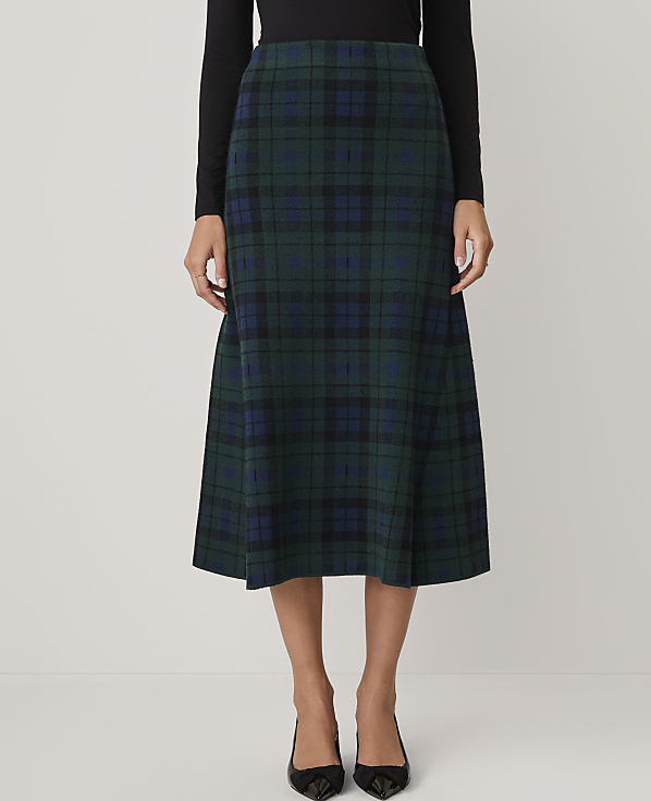 A person wears a black top and a green and blue plaid midi skirt against a plain white background.