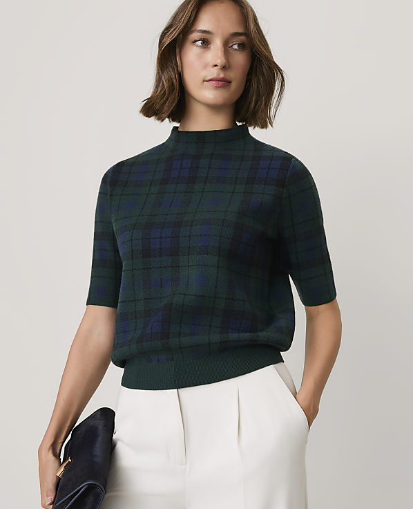 A woman wearing a dark green plaid short-sleeved sweater and white trousers stands against a plain, light-colored background.
