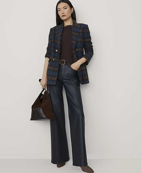A woman wearing a plaid blazer, dark top, and wide-leg faux leather trousers stands against a plain, light-colored background.