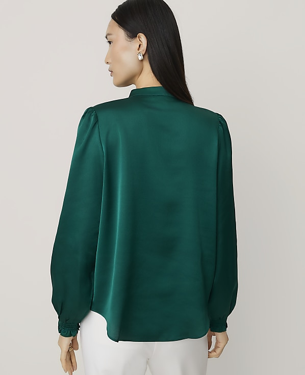 A woman wearing a dark green satin blouse and white trousers stands against a plain, light-colored background.