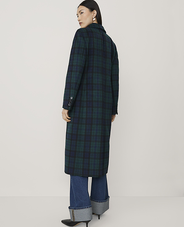 A woman wearing a long green and blue plaid coat and cuffed blue jeans stands against a plain, light-colored background.