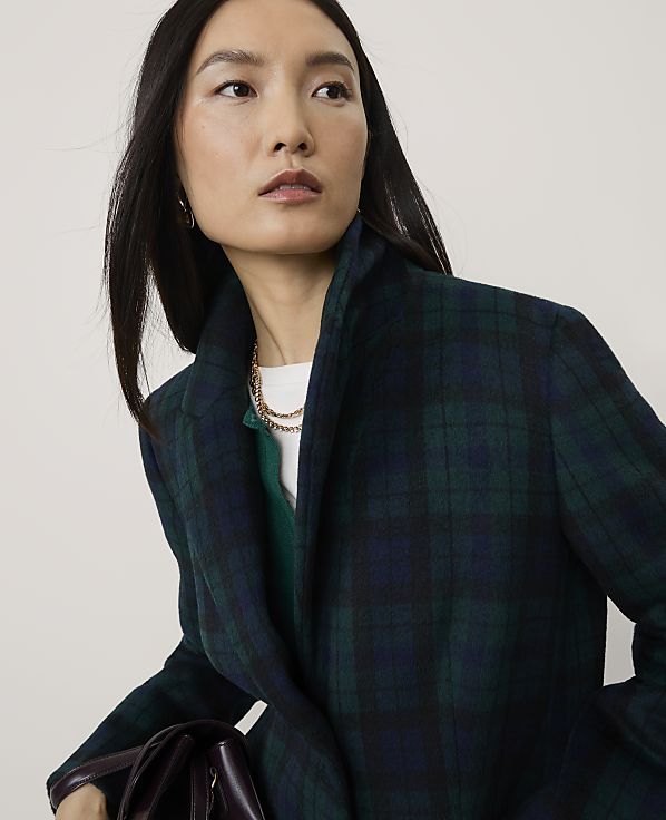 A woman wearing a dark green plaid blazer and a gold chain necklace poses against a plain, light-colored background.