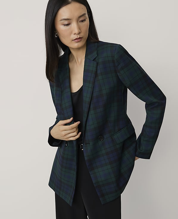 A woman wearing a dark green plaid blazer and black trousers stands against a plain, light-colored background.
